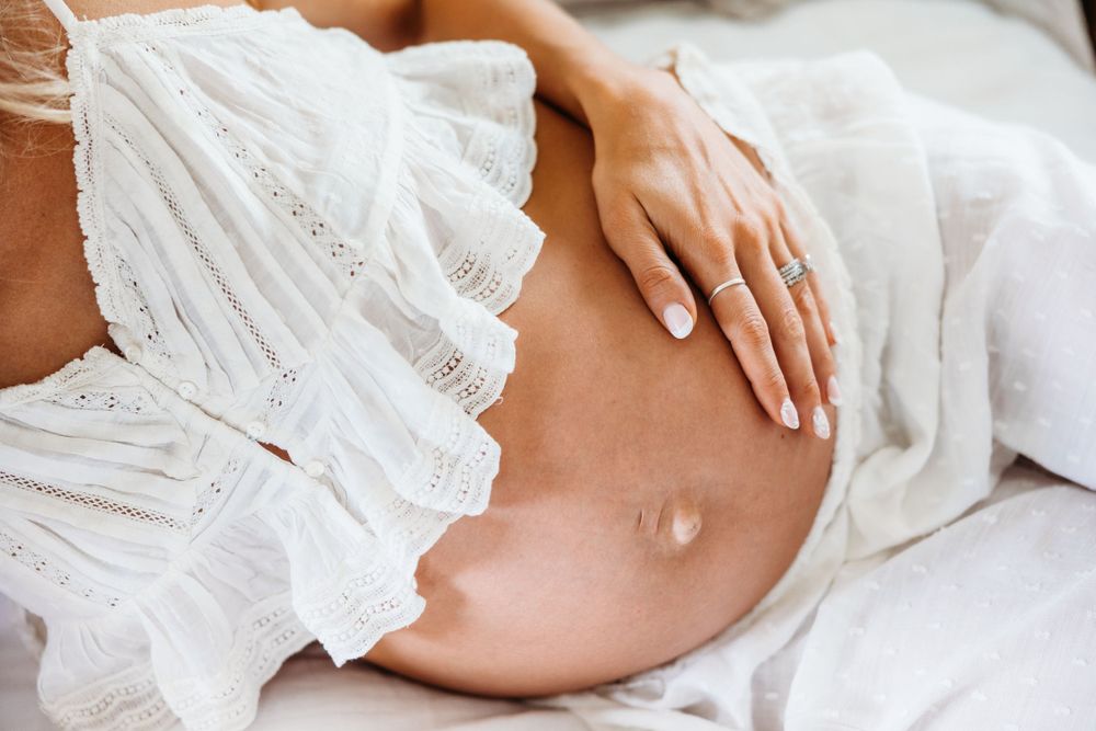 Close-up of pregnant belly in white lace dress against white bedding.