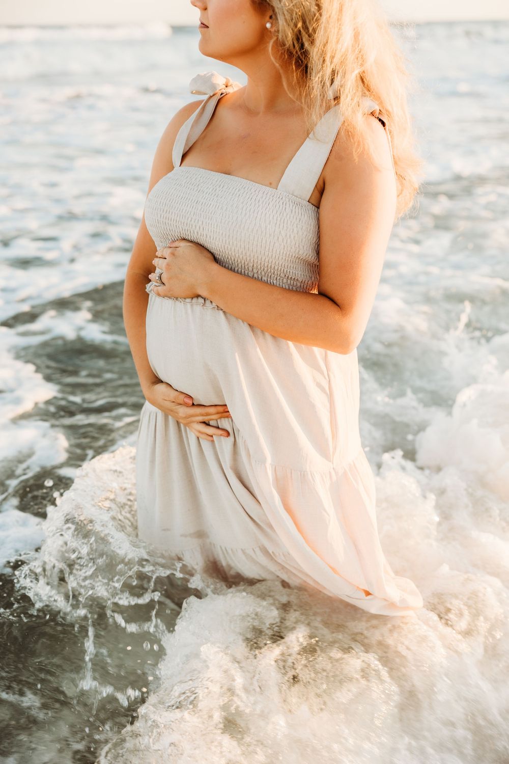 A pregnant person in a white dress stands in ocean waves at sunset holding their baby bump.