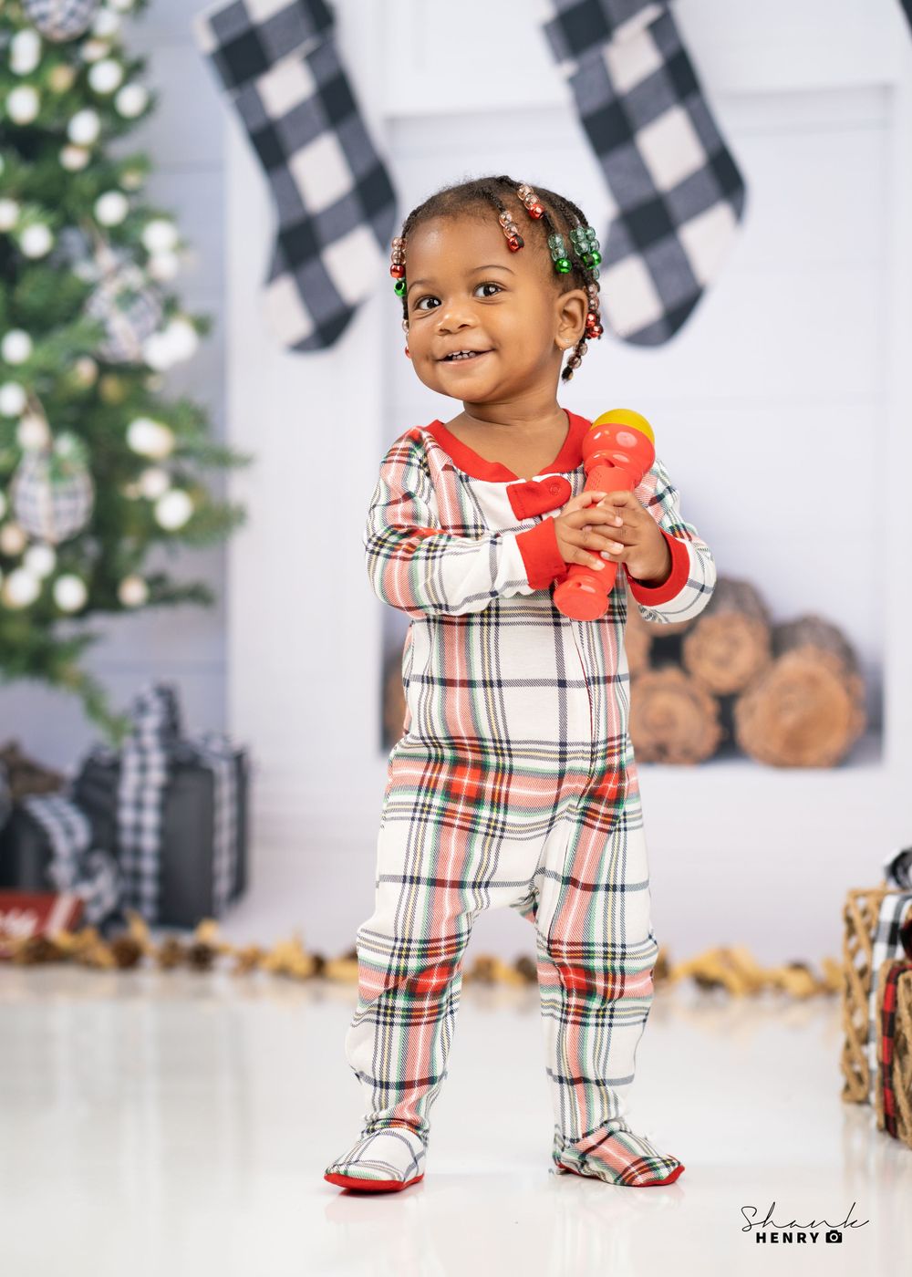 Toddler in plaid holiday pajamas holds red toy in front of Christmas decorations.