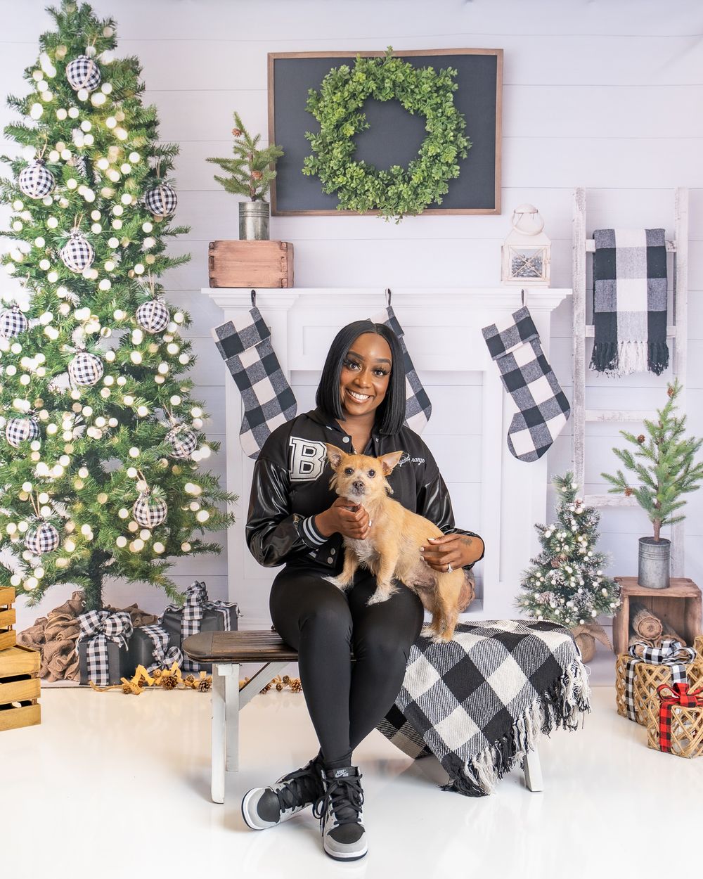 Woman in black jacket holds small brown dog in front of Christmas tree and buffalo check stockings.