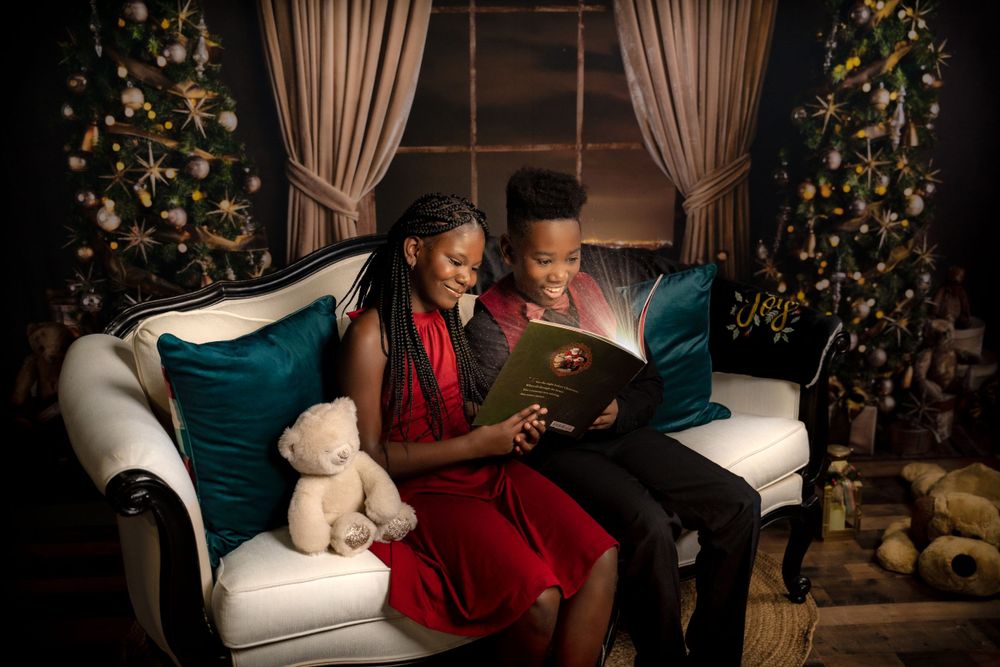 Two people sit cozy on white couch reading book together by decorated Christmas trees.