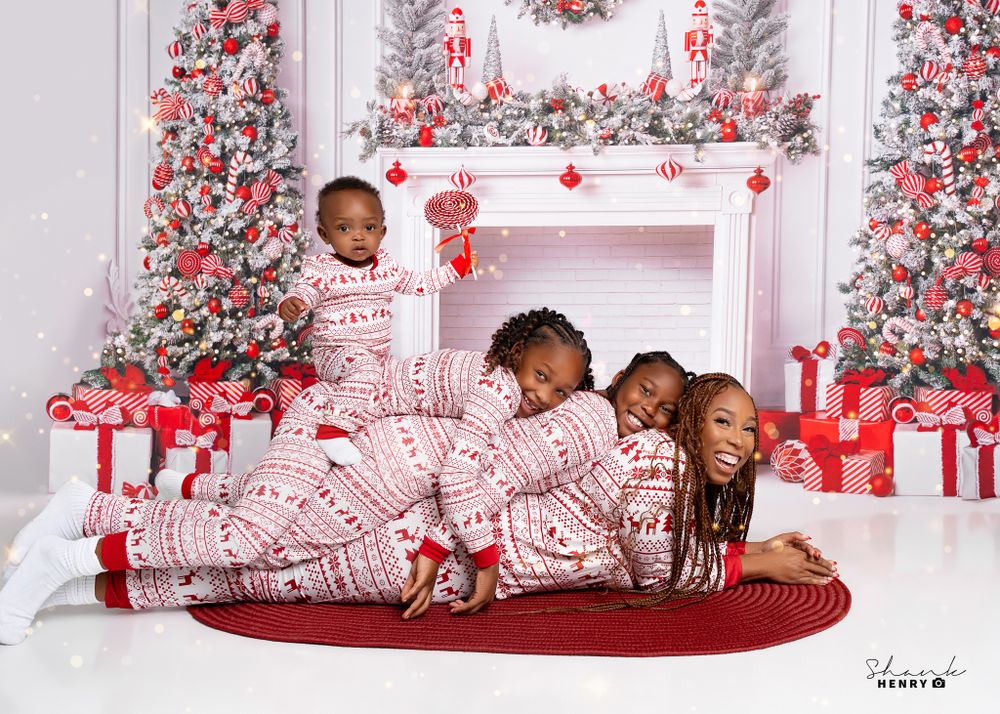 Family lying together in matching Christmas pajamas by decorated fireplace with wrapped presents.