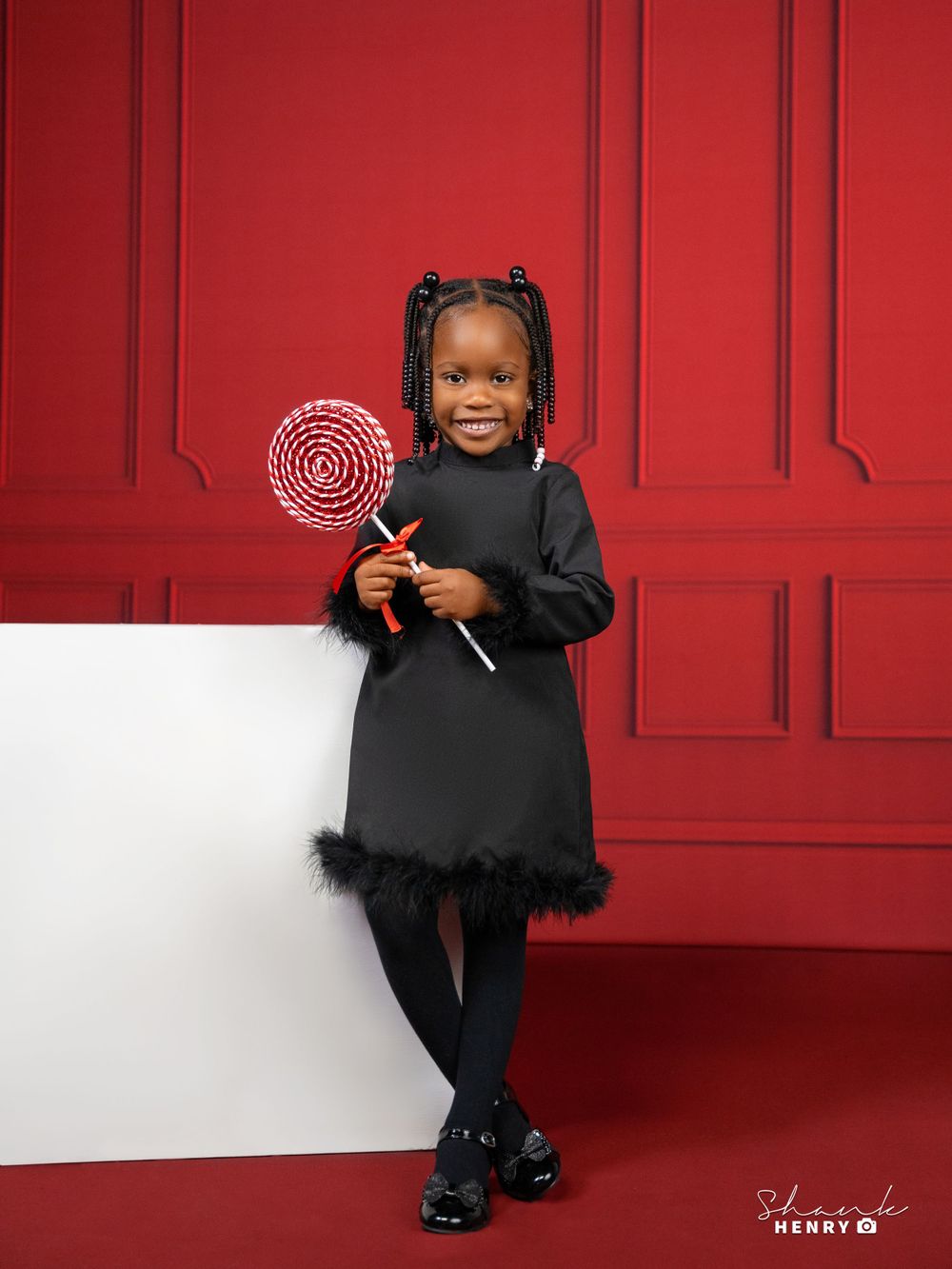 A cheerful child in a black dress holds a red and white lollipop against a bright red wall backdrop.