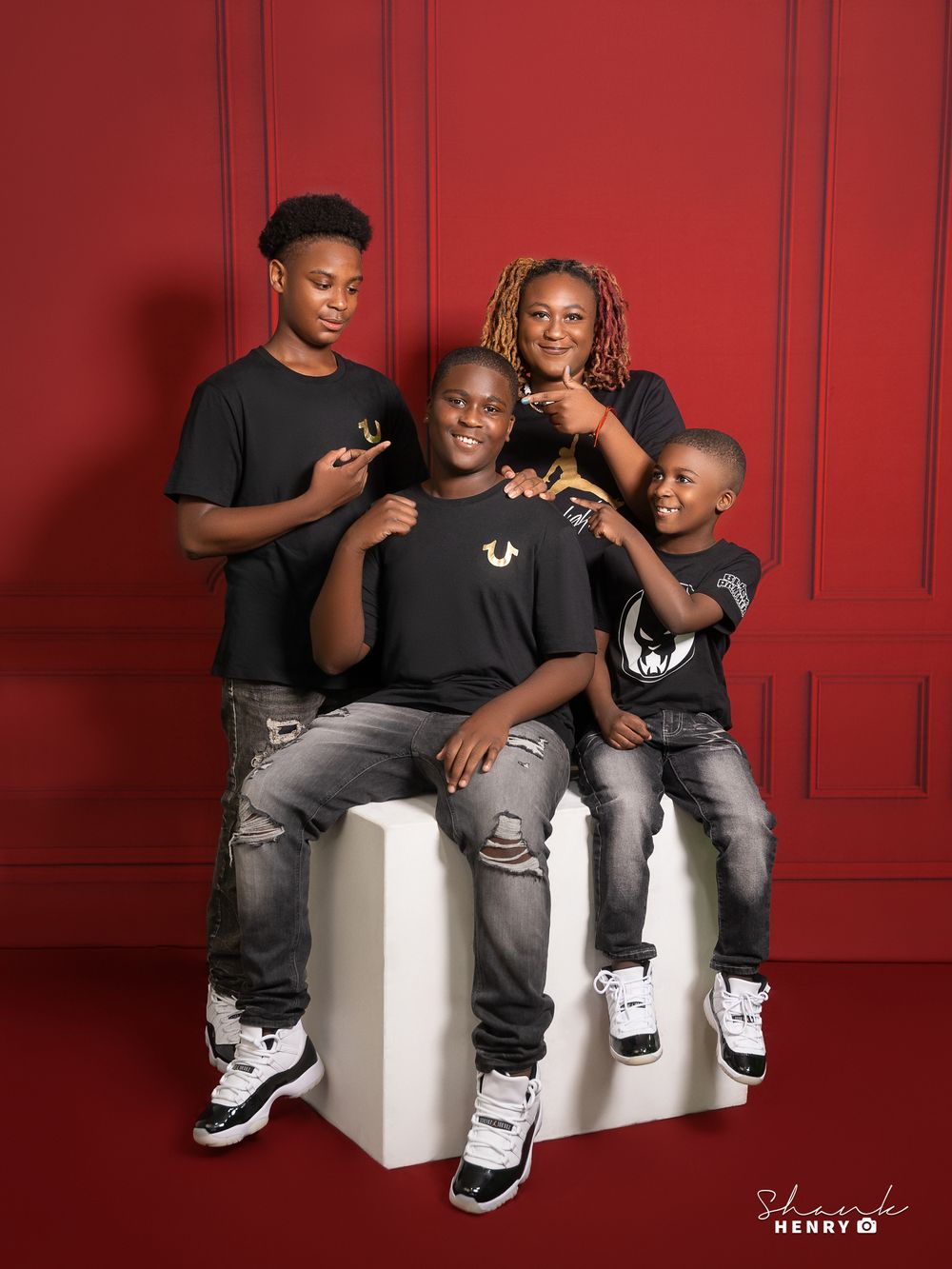 Group photo in black t-shirts and distressed jeans posed on white blocks against red wall.