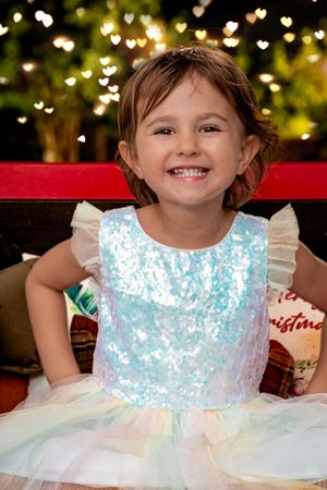 Young child wearing a sparkly blue and white tulle dress smiles in front of Christmas lights.