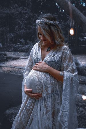 Delicate white lace maternity gown photographed in moody evening light with bokeh effects.