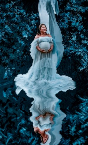 Flowing blue dress creates ethereal effect against dark foliage in maternity portrait.