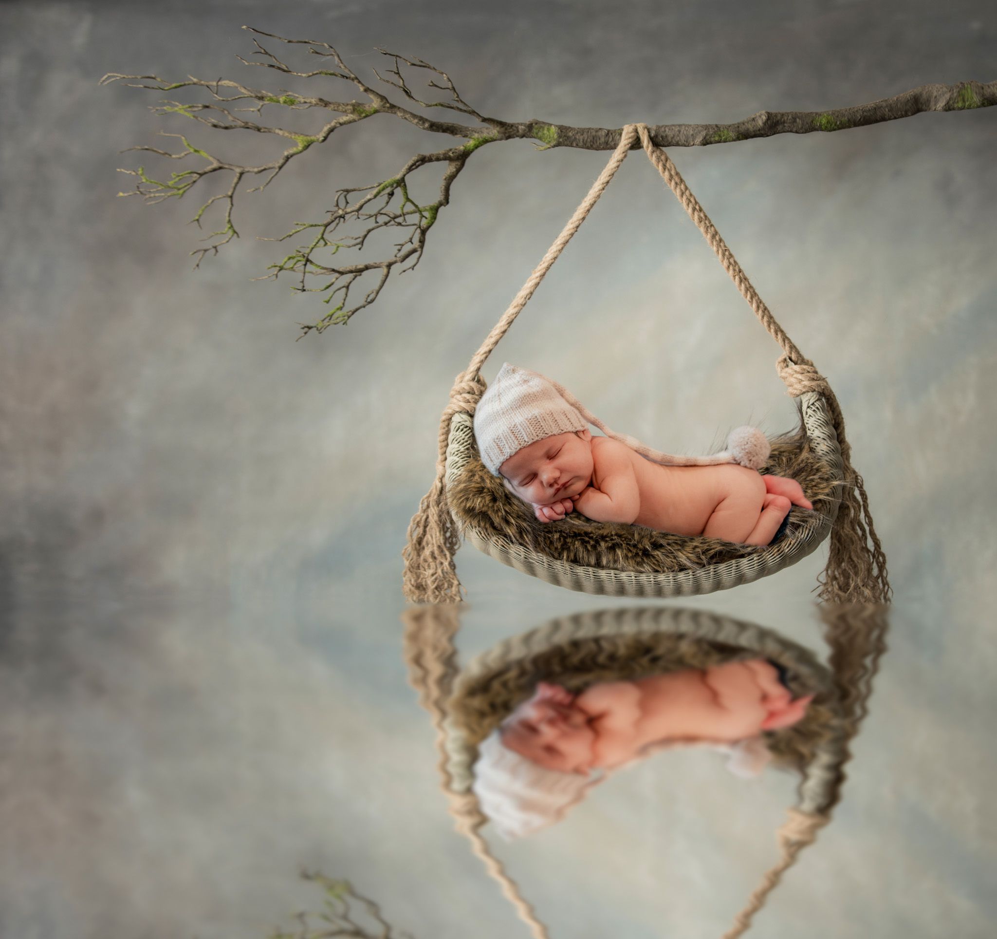 Newborn baby sleeping peacefully in a hanging knitted hammock against a grey backdrop.