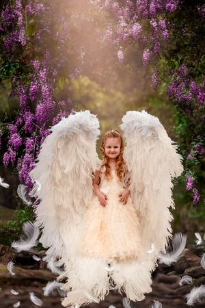 White feathered angel wings glow against purple wisteria flowers in dreamy garden setting.