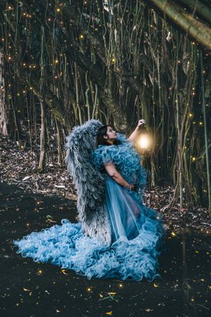 Dramatic blue dress and angel wings photographed in moody forest setting with glowing lights.
