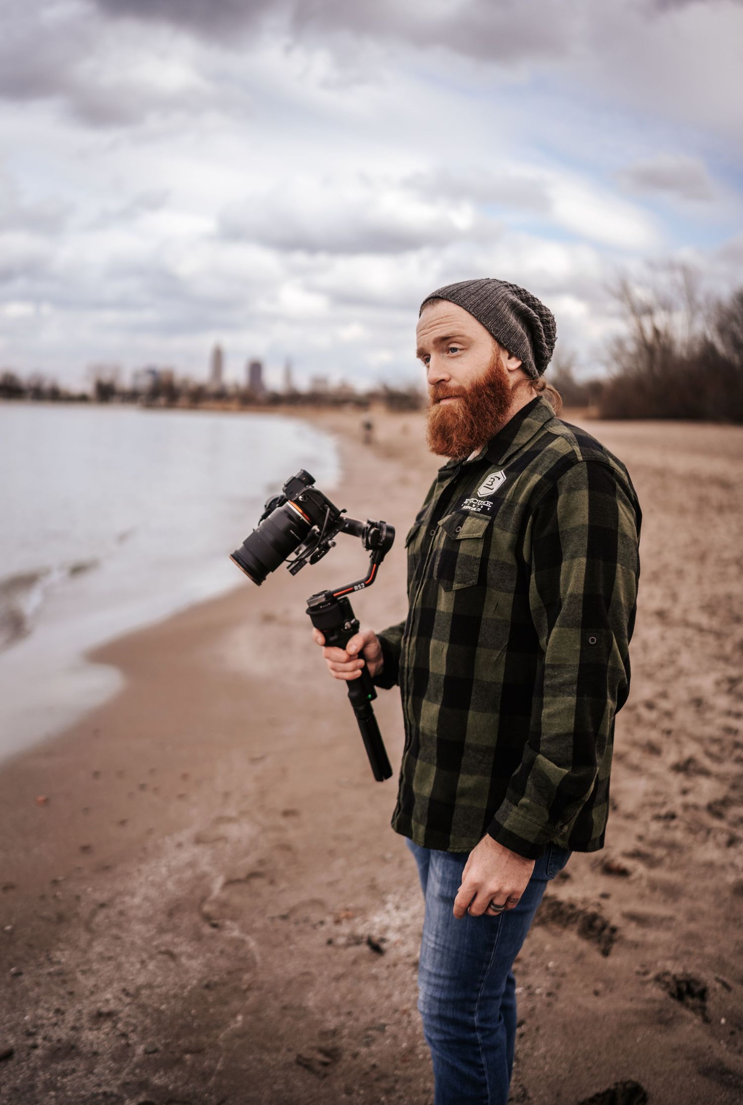 Photographer with red beard wearing plaid and beanie holding camera gear on beach.