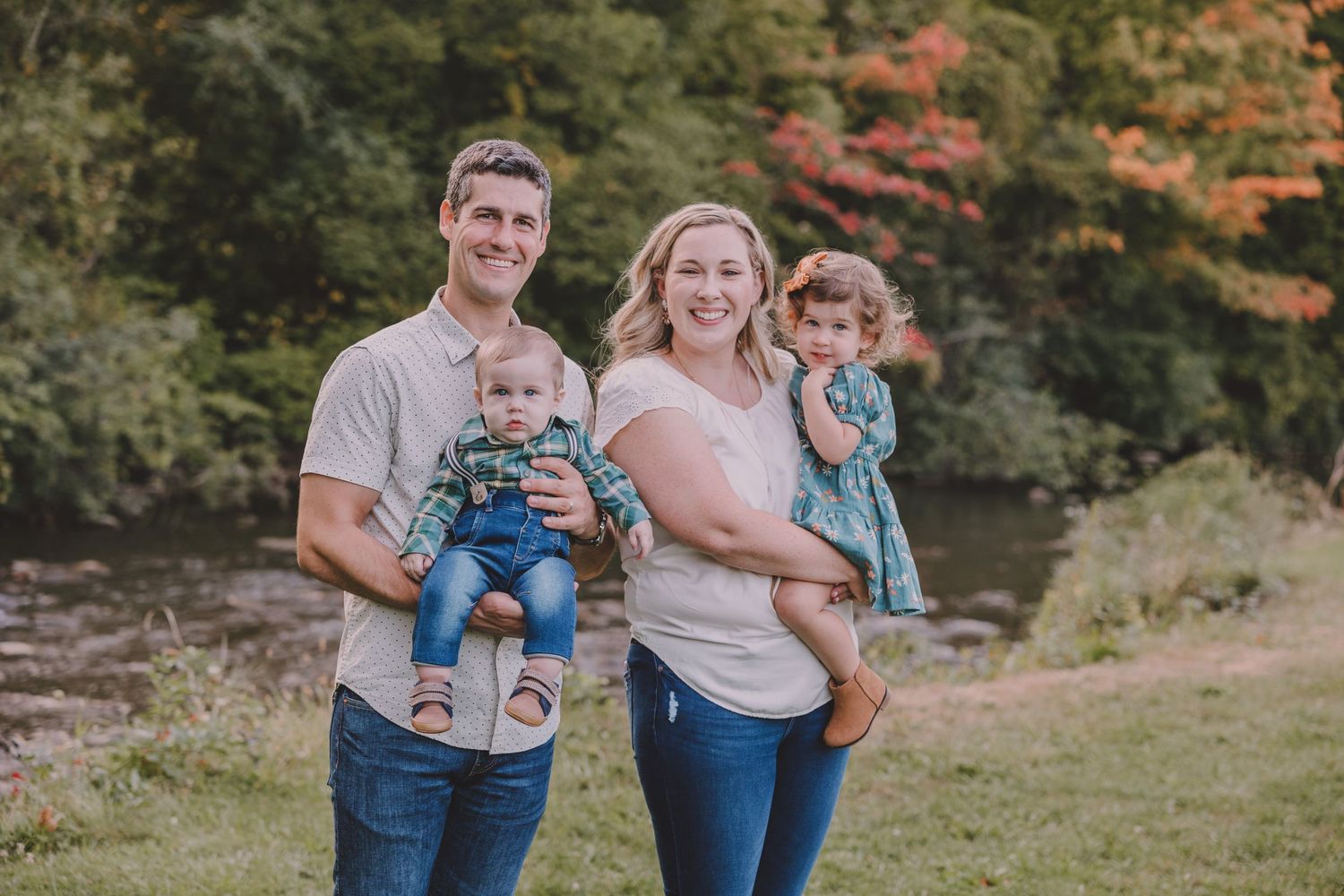 Family portrait of four members standing together on a grassy slope with autumn scenery behind them.