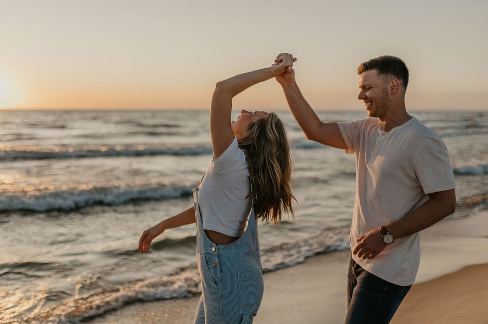 Couple dancing and twirling together on beach at sunset with ocean waves in background.