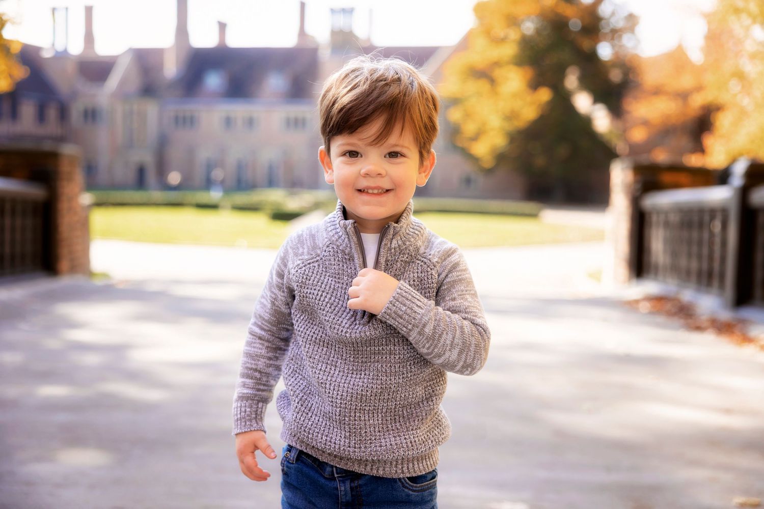 Young child in gray sweater smiling outdoors with autumn manor house in background.