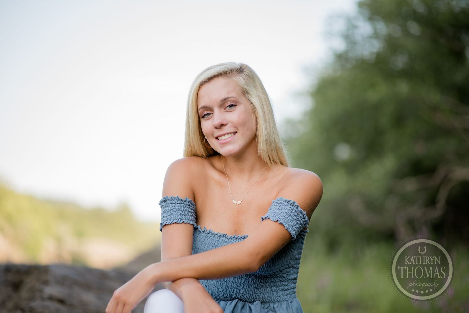 Woman in blue smocked top sits casually on rocks during outdoor photo session.