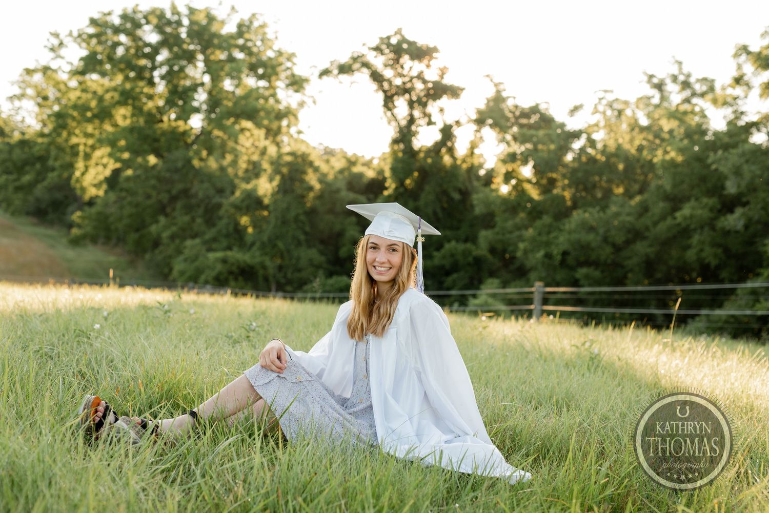 Graduate in traditional white regalia sits gracefully in a sunlit meadow with trees in background.