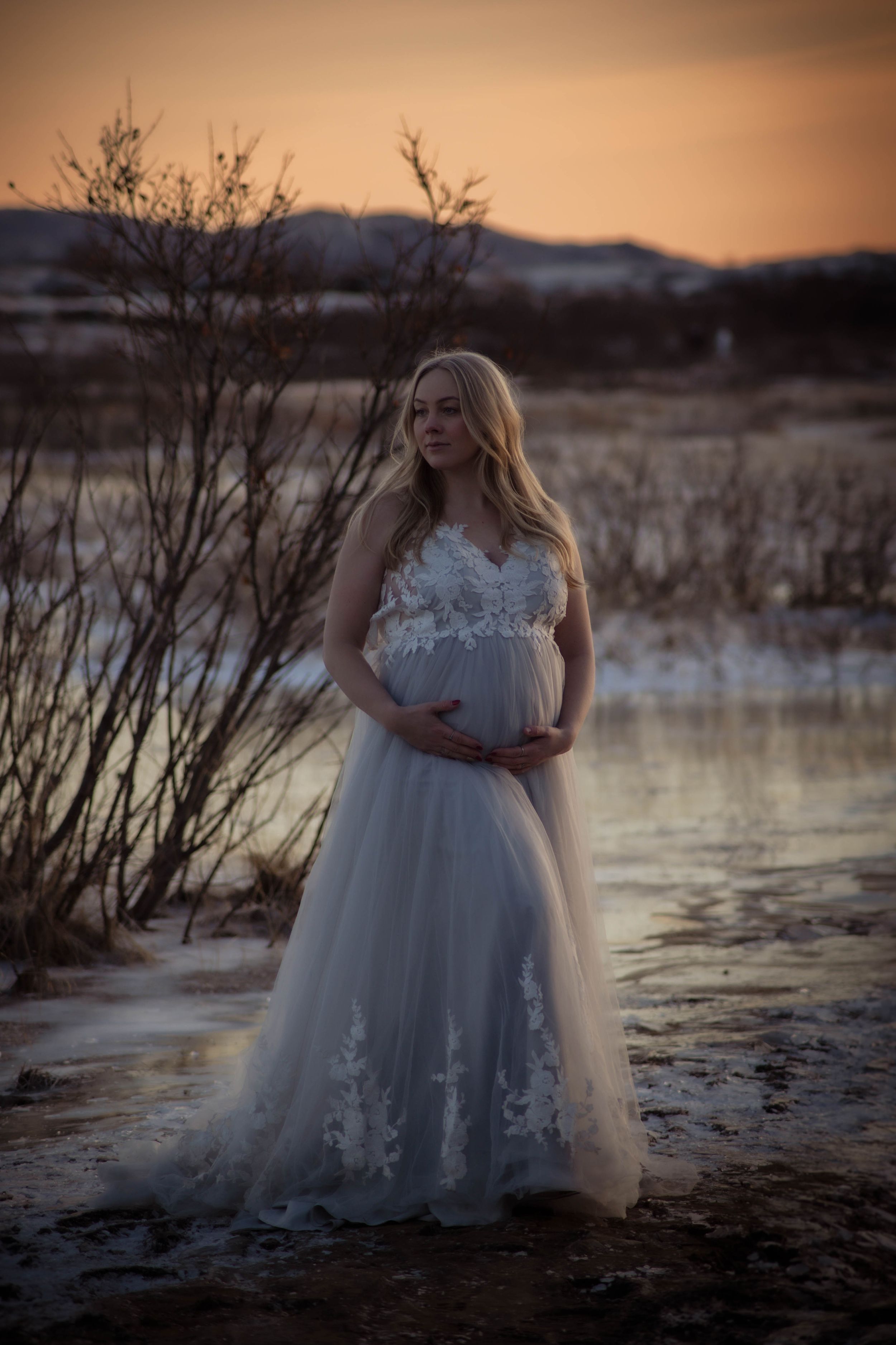 Pregnant woman in elegant white lace tulle gown standing near a snowy lake at sunset.