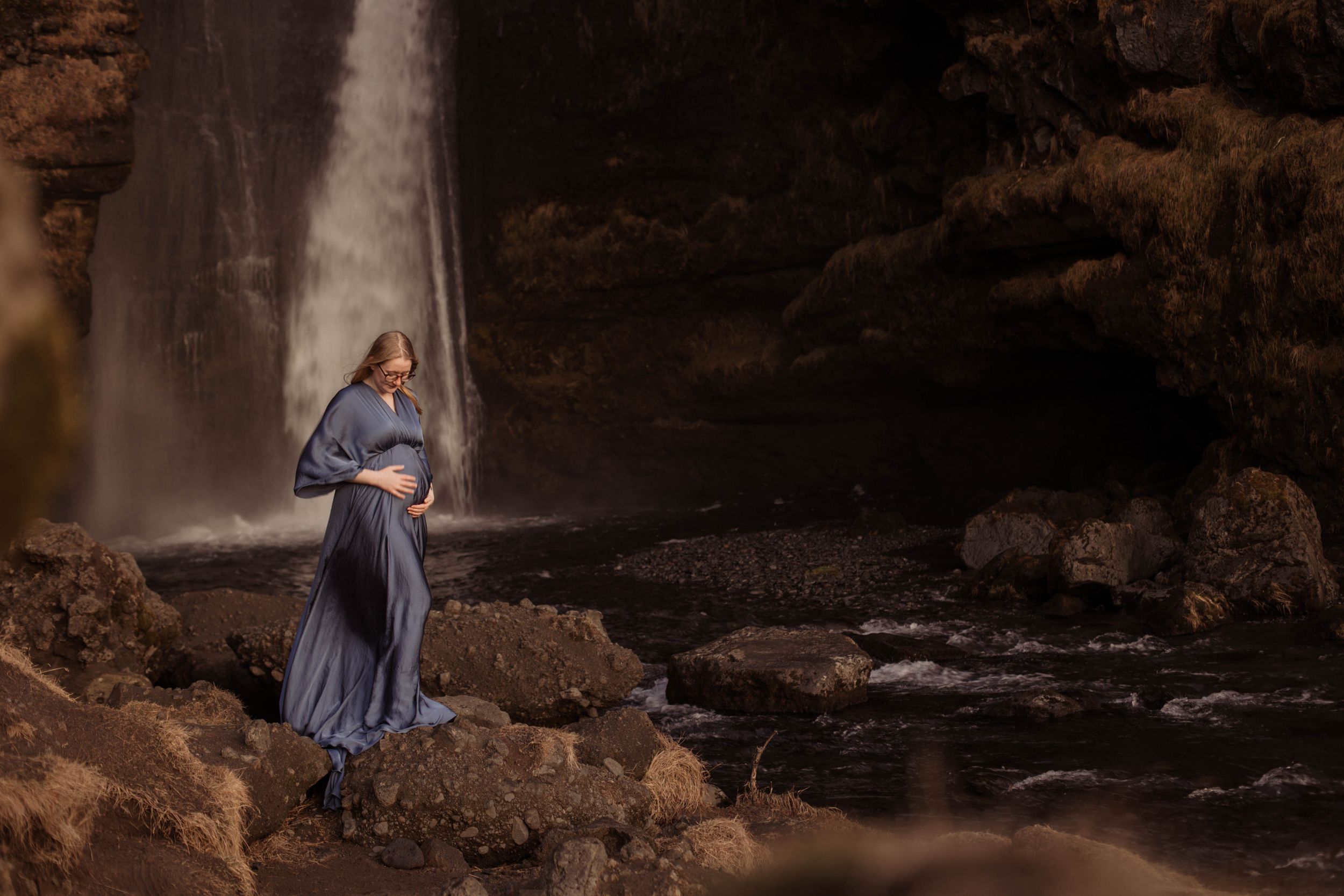 Pregnant woman in blue maxi dress standing on rocks before a dramatic waterfall backdrop.