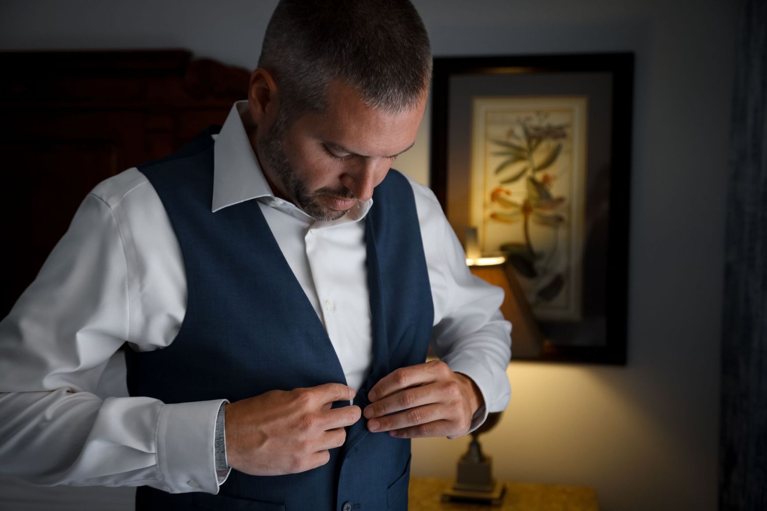Man in navy vest and white shirt buttoning up in dimly lit room.