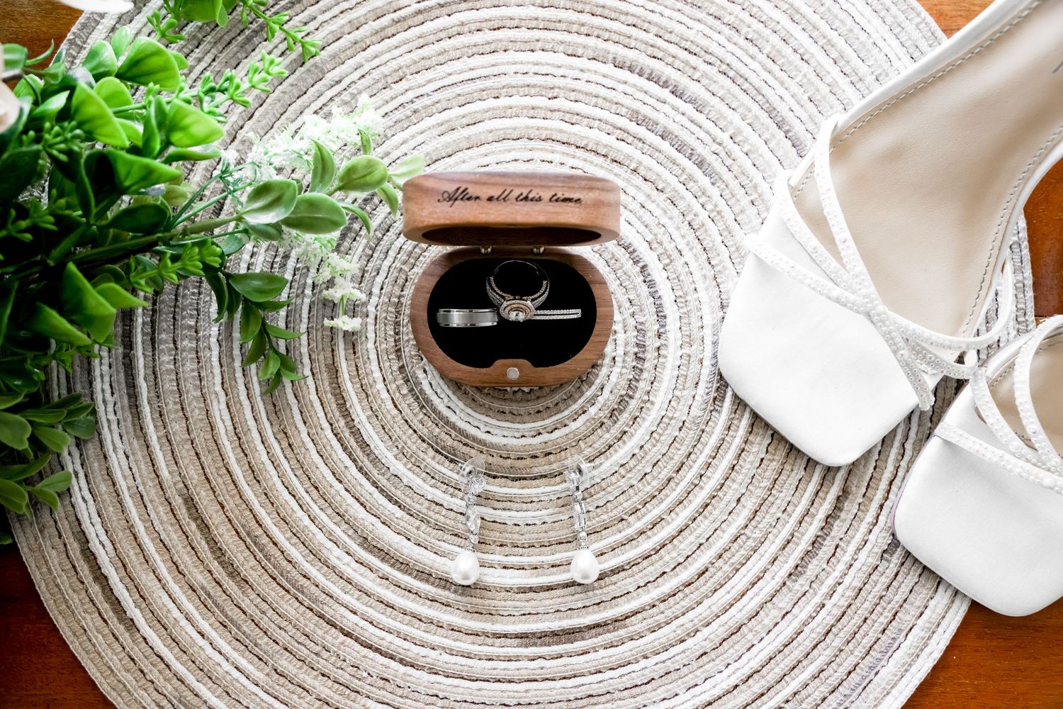 Wedding rings displayed on a circular textured placemat with bridal shoes and greenery.