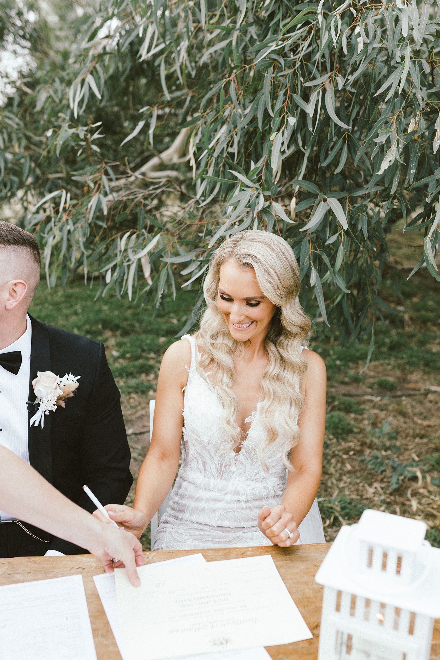 Newlyweds signing their marriage license at a wooden table under eucalyptus trees.