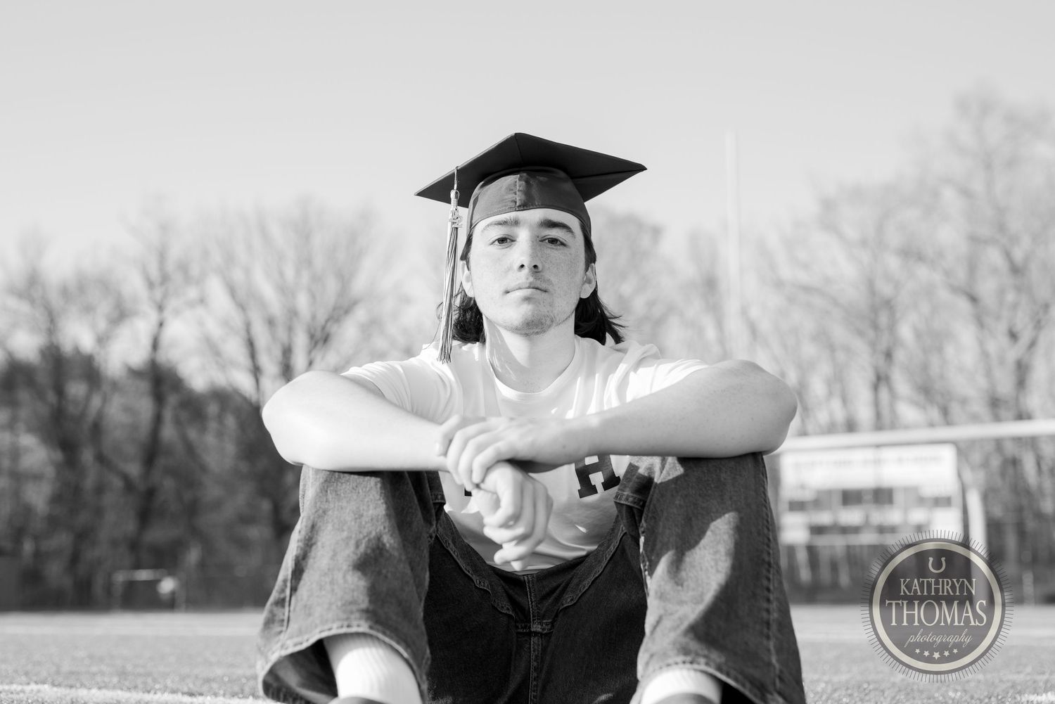 Black and white portrait of graduate sitting on field in academic regalia.