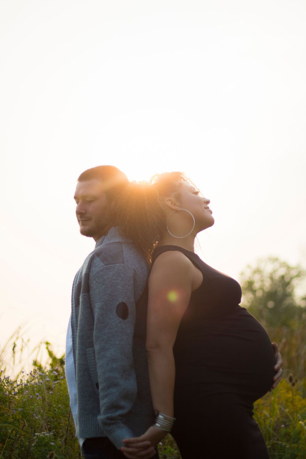 A couple stands back-to-back in a field during golden hour sunset.