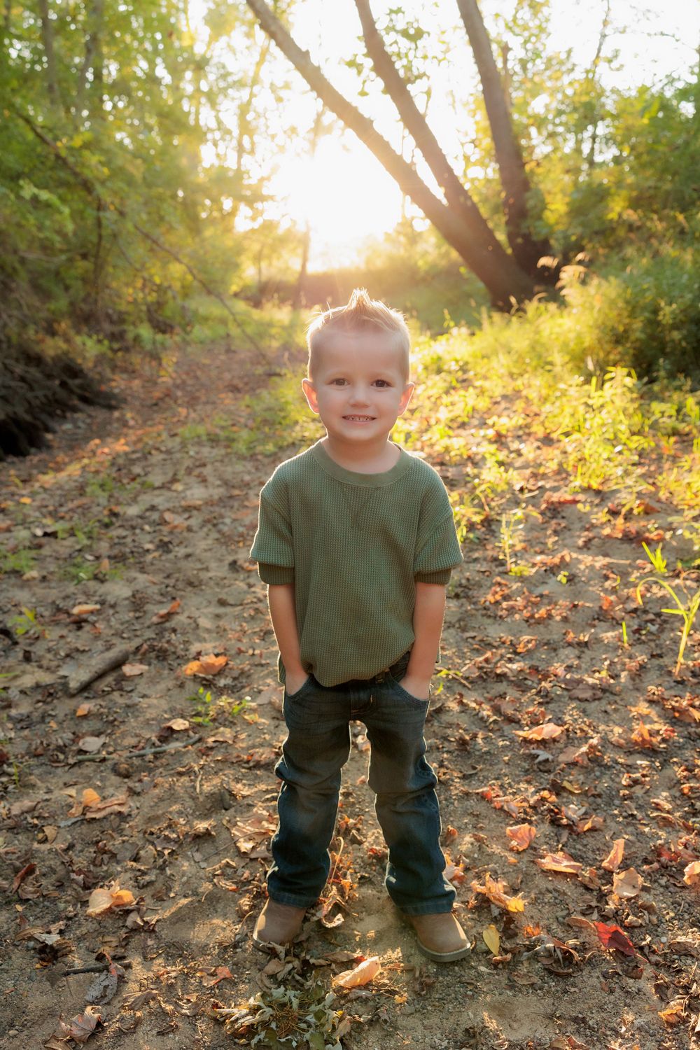 Child in green shirt and jeans stands on leaf-covered forest path in golden sunlight.