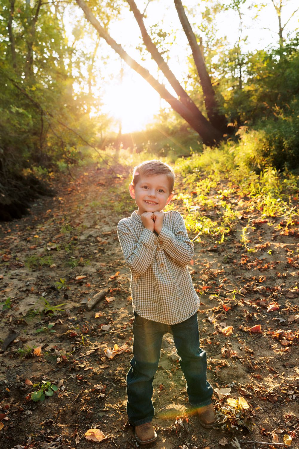 Child in checkered shirt poses playfully on forest trail during sunset.