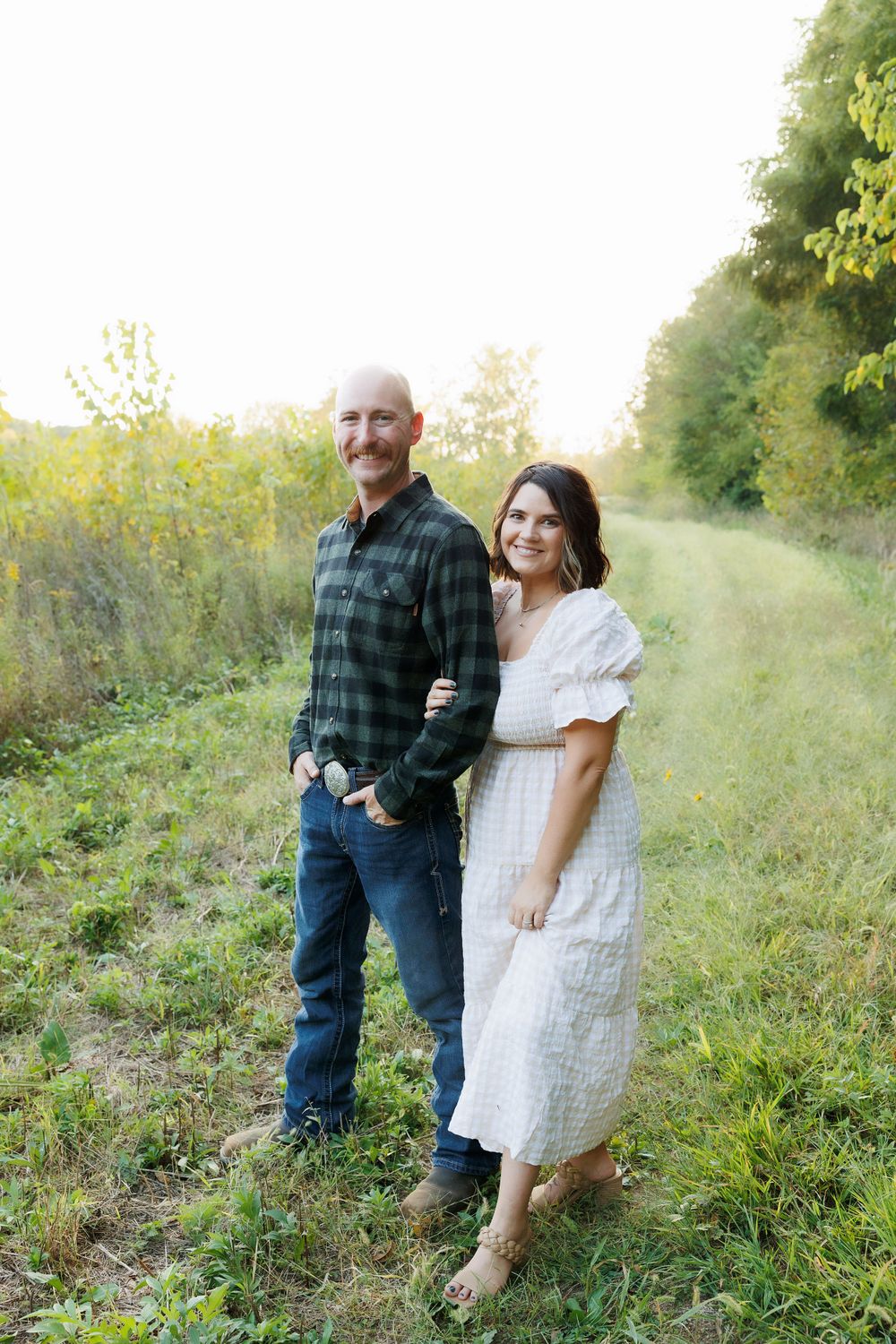 Couple poses together on a grassy trail wearing green plaid and white dress.