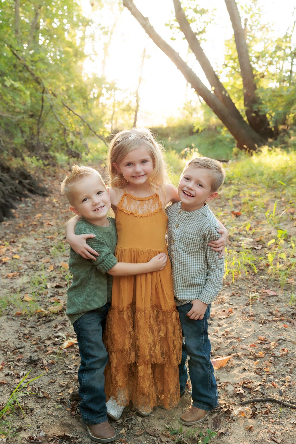 Three children embrace and laugh together on a sunlit autumn path surrounded by fallen leaves.