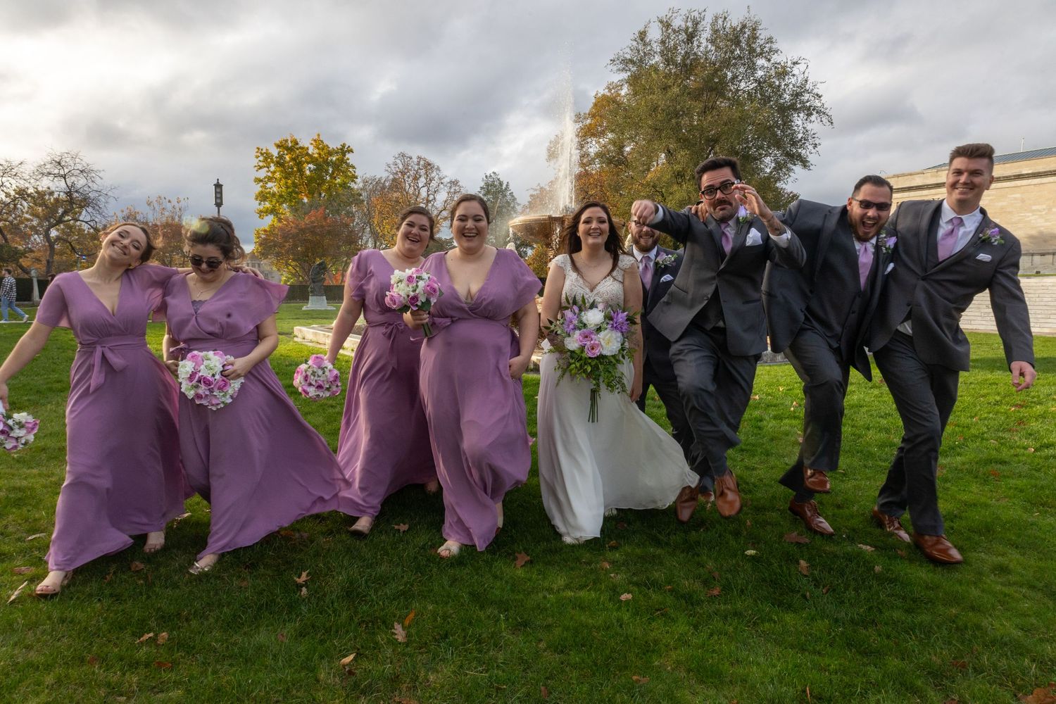 Wedding party in purple dresses and gray suits poses together on grassy hillside.