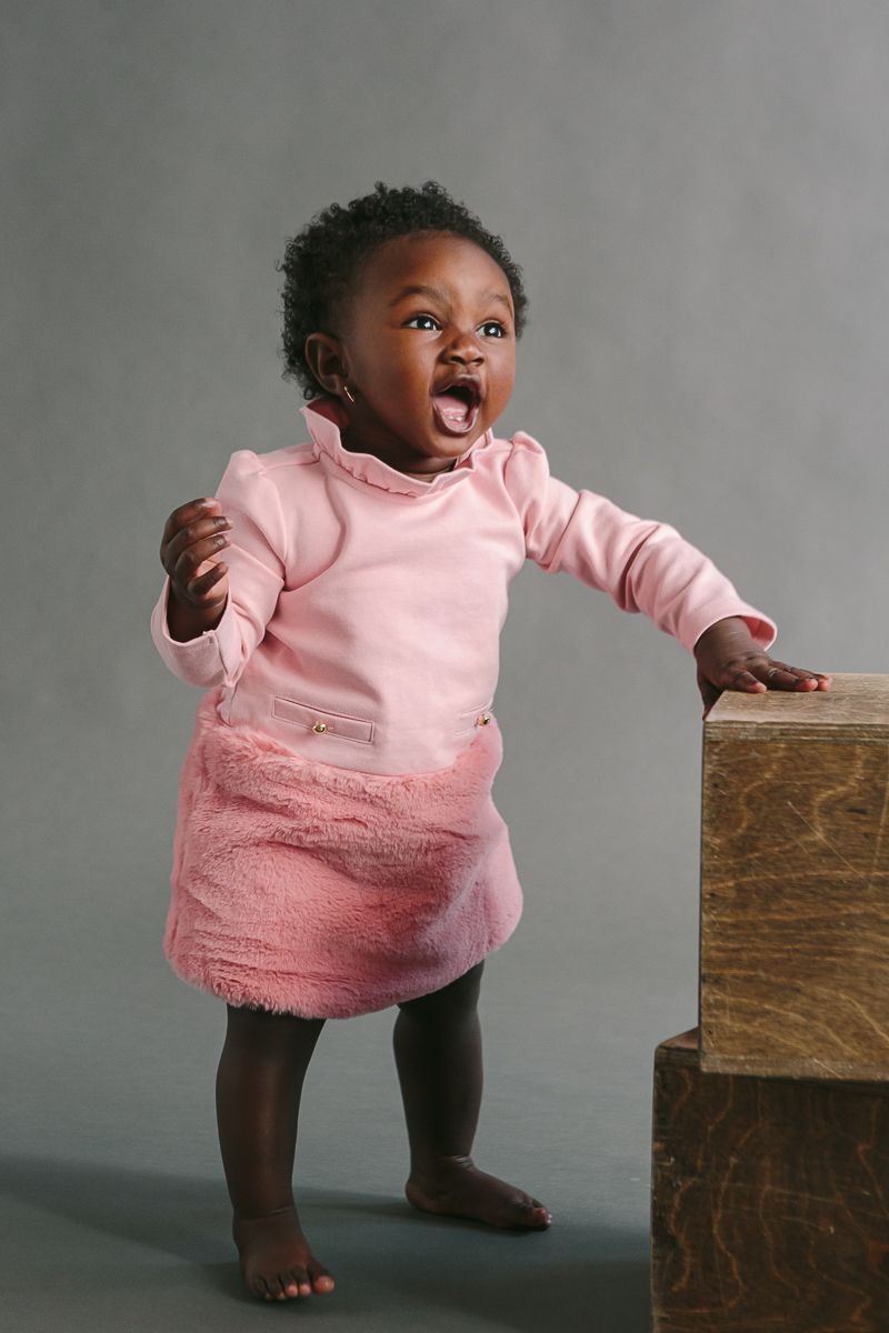 a child on a pink dress stands against a grey backdrop during a studio session by NYC Family Photographer Paula Ortiz.