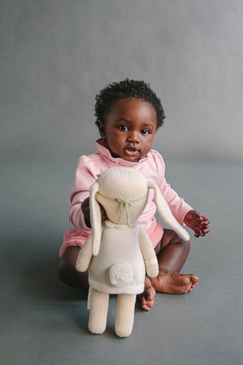 a child on a pink dress is sitting and holding a cuddle and kind bunny toy. The child is sitting on a grey backdrop