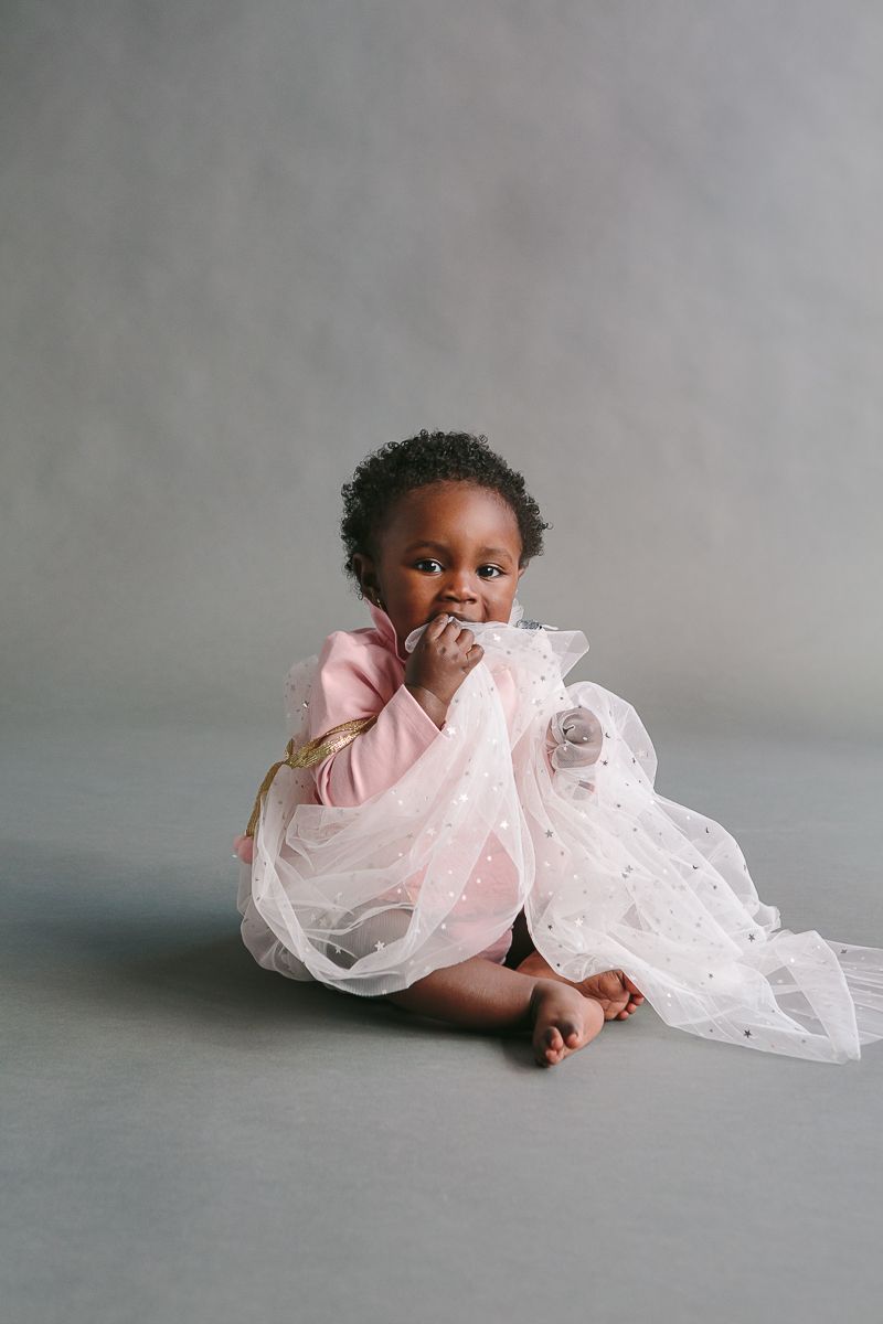 a child on a pink dress sits and eats a white cape during a studio session by NYC Family Photographer Paula Ortiz