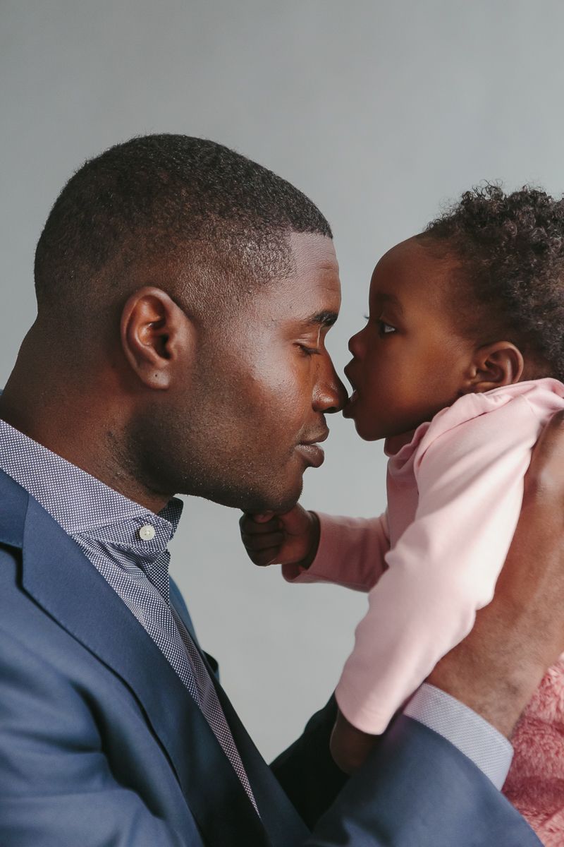 a black father wearing a blue suit closes his eyes while his daughter in a pink dress bites his nose on a grey studio