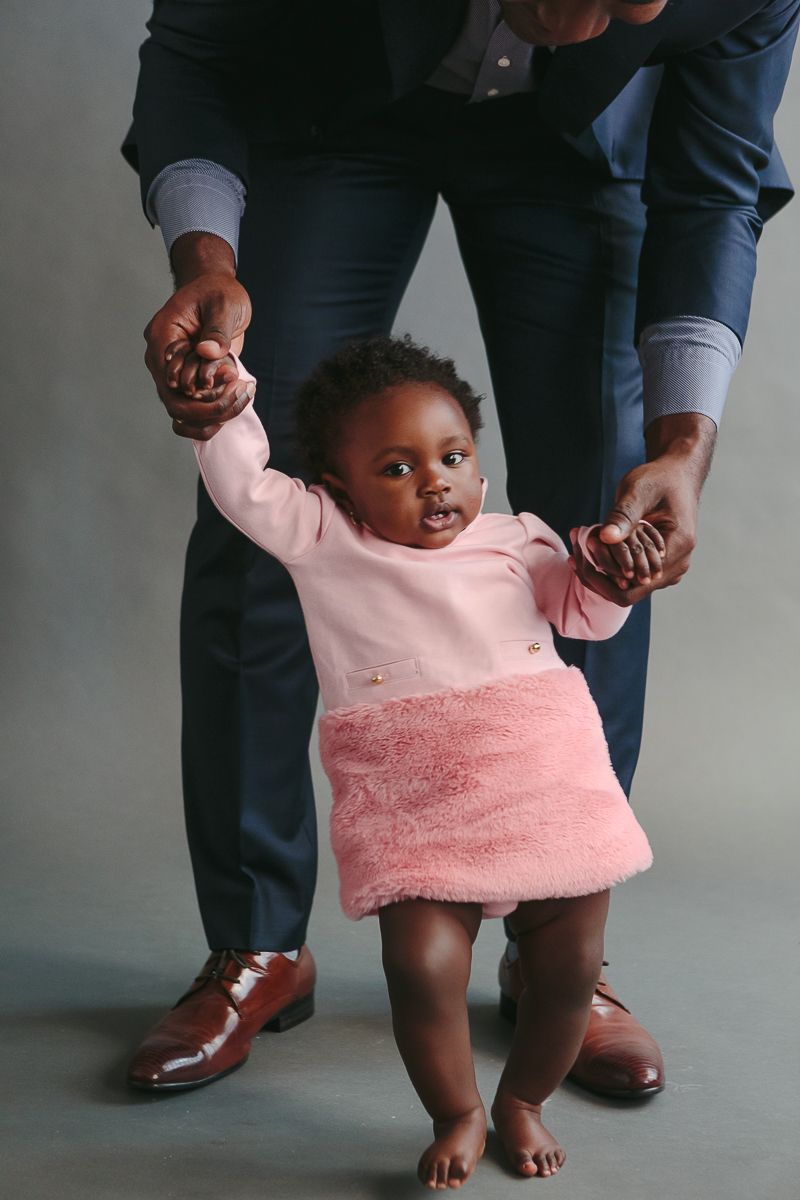 a little girl on a pink dress stands with the help of her dad not fully pictured in the background during a NYC studio