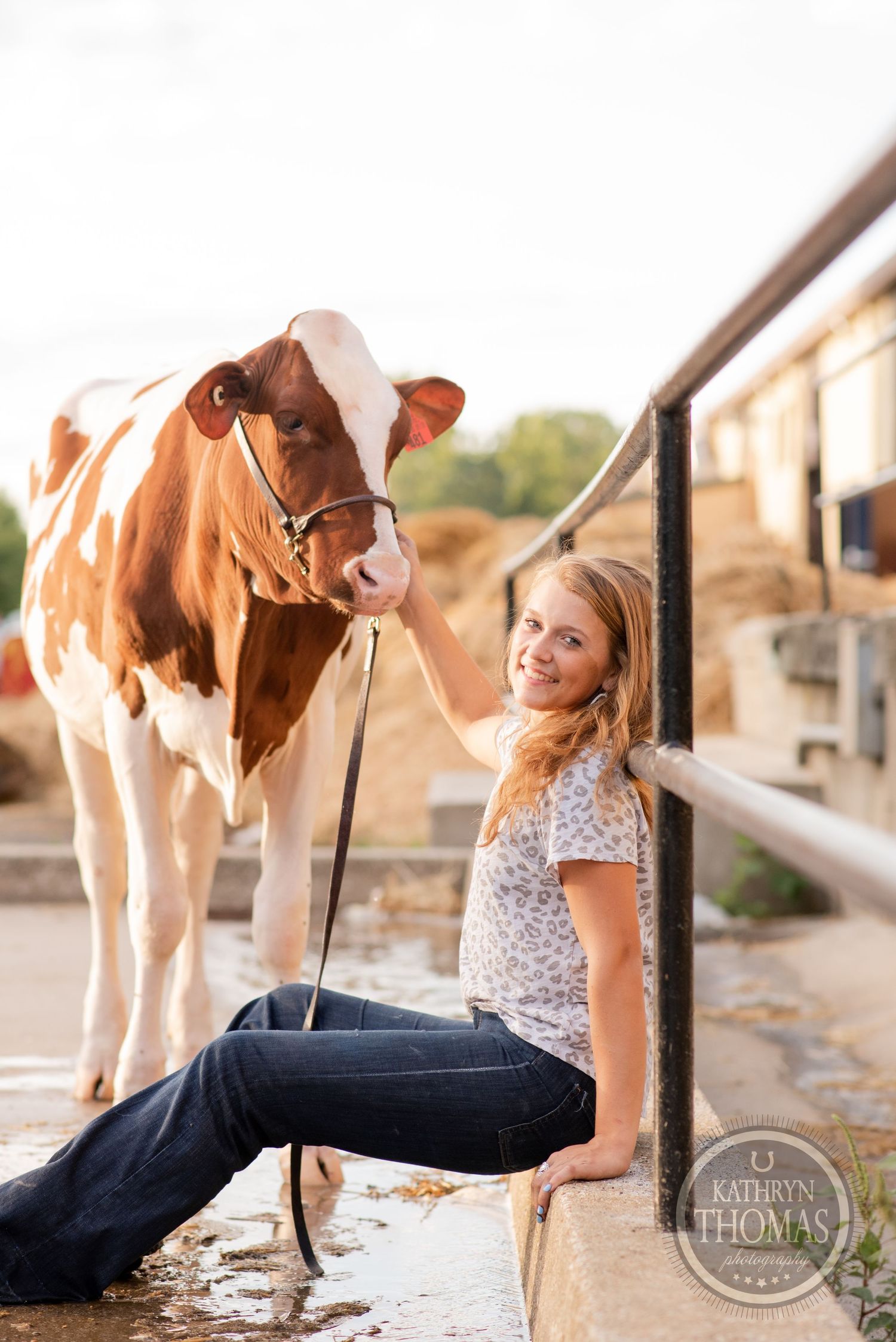 A brown and white cow stands next to a person leaning against a railing in casual farm attire.