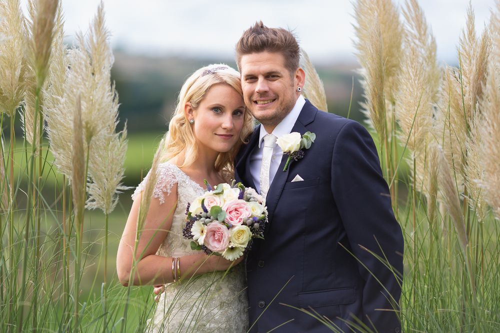 A bride with her bouquet and and groom stand close together amongst reeds and tall grass, looking at the camera.