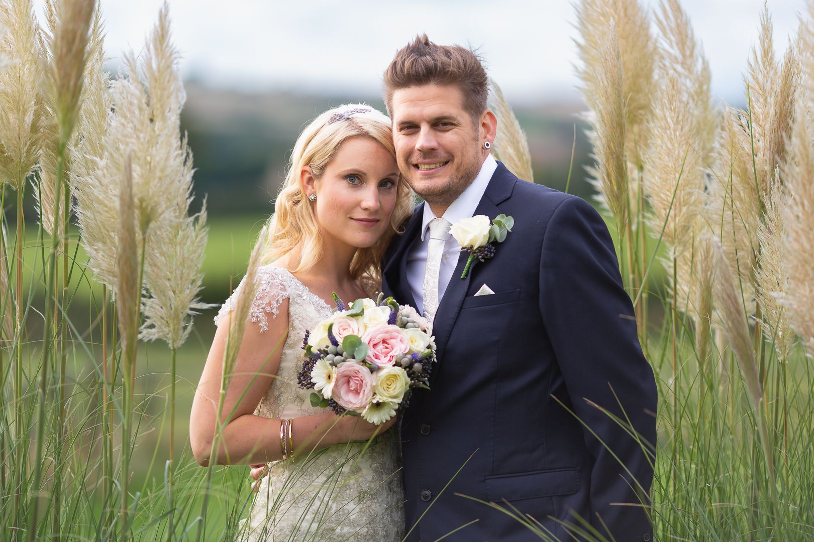 A bride with her bouquet and and groom stand close together amongst reeds and tall grass, looking at the camera.