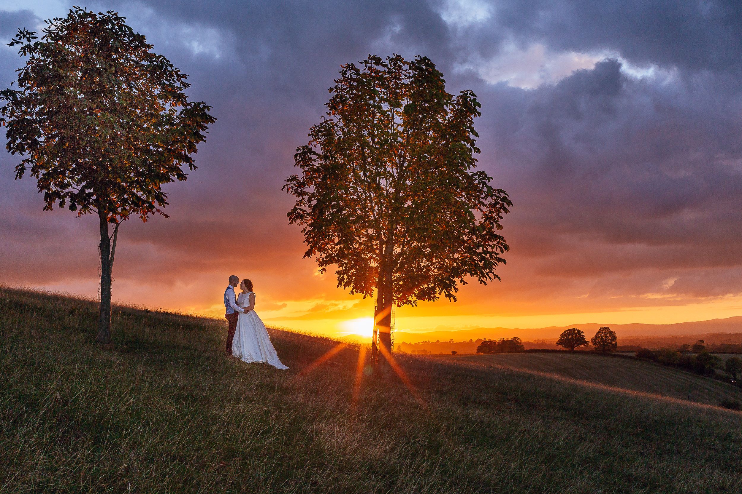 A bride and groom stand between two trees on a hillside as the sun sets against an orange and purple sky.
