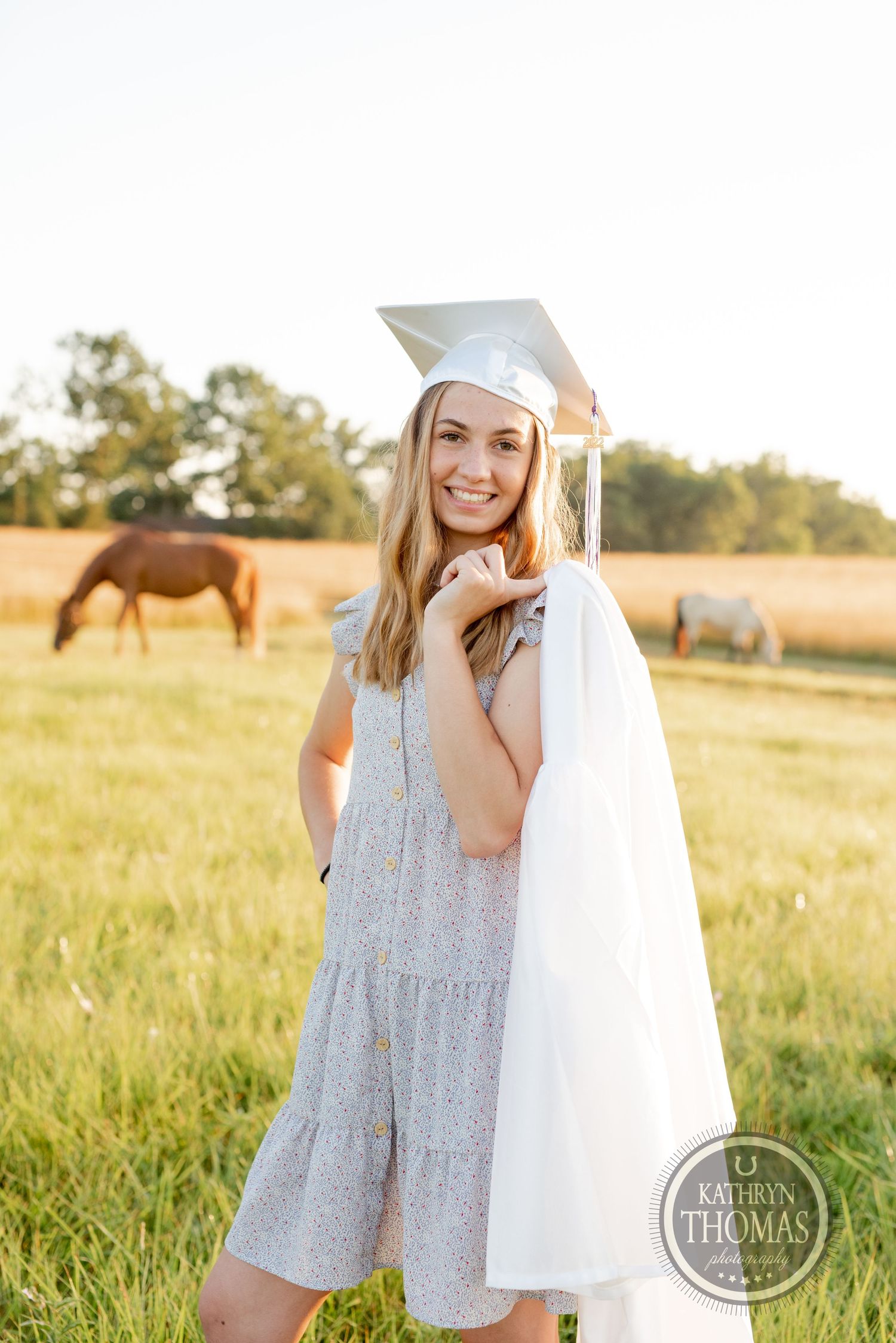 Graduate wearing white gown and cap poses in a sunlit meadow with horses grazing behind.