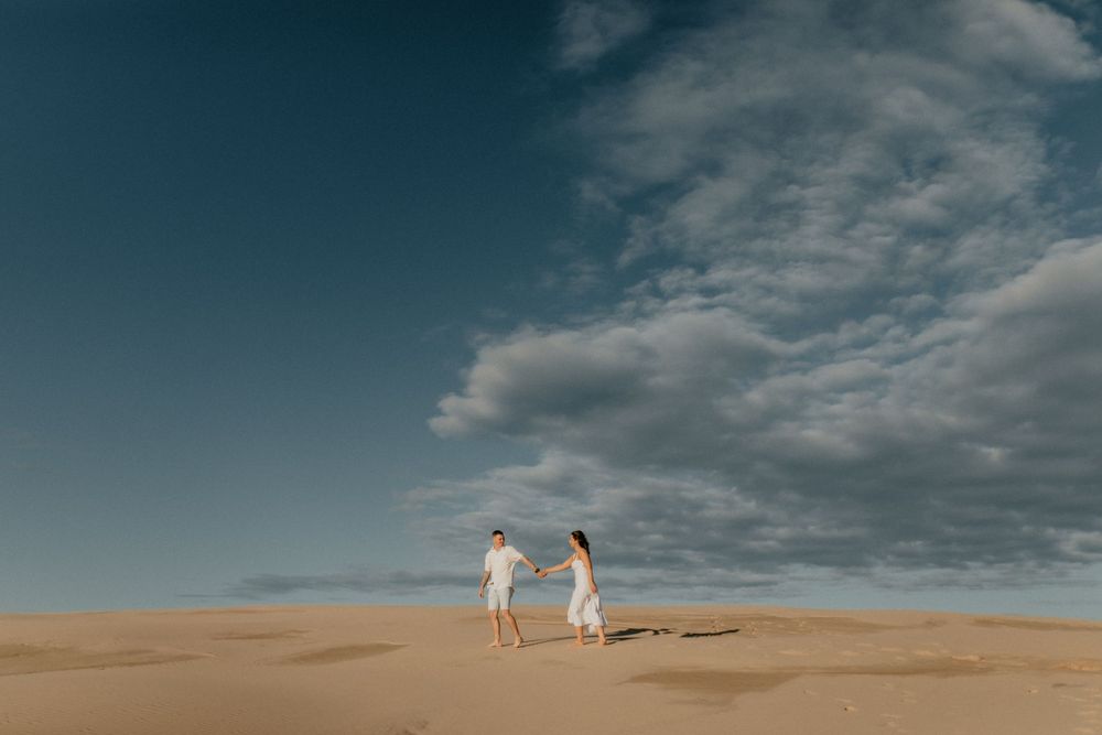 Couple holding hands while walking across sand dunes under a dramatic blue sky.
