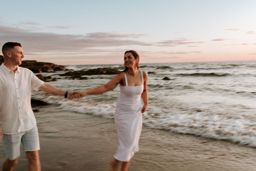Couple walking along beach shoreline during sunset wearing white outfits.