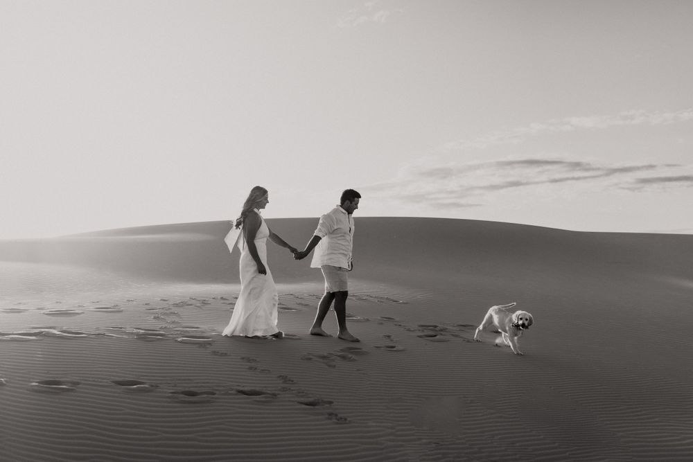 A couple walks along desert sand dunes with their dog trailing behind.