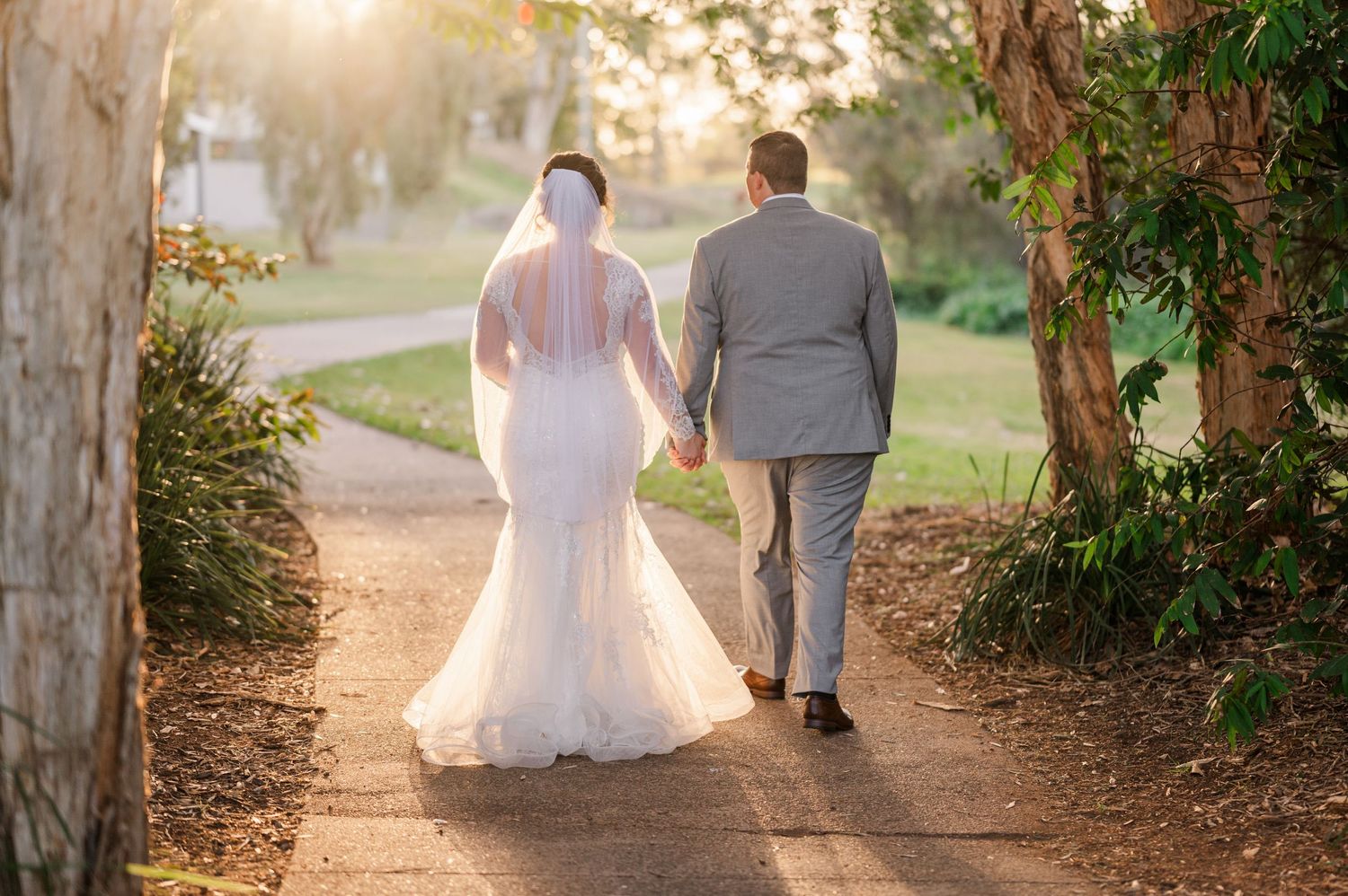 A couple walking hand in hand down a tree-lined path at sunset in their wedding attire.