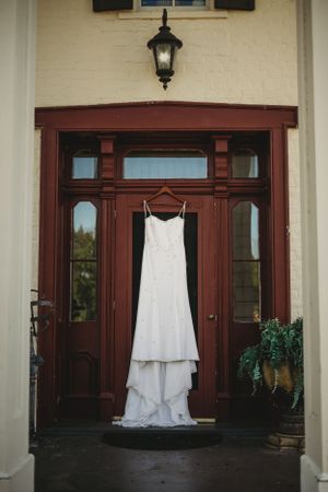 White wedding dress hanging on red wooden doors of elegant building entrance.
