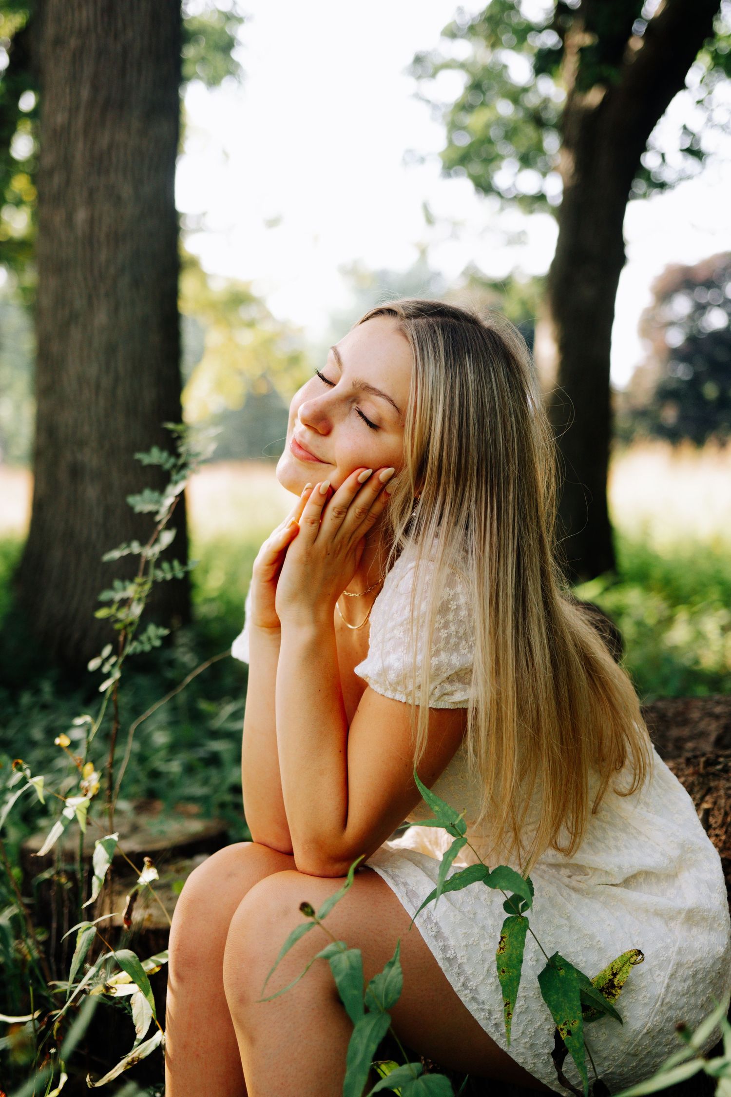 A dreamy outdoor portrait of a woman in a white dress sitting among greenery with closed eyes and serene expression.