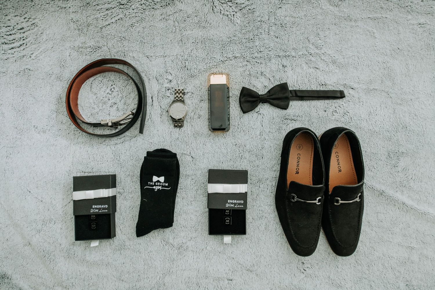 Groom accessories including black loafers, bow tie, belt, socks and watch laid out on gray surface.
