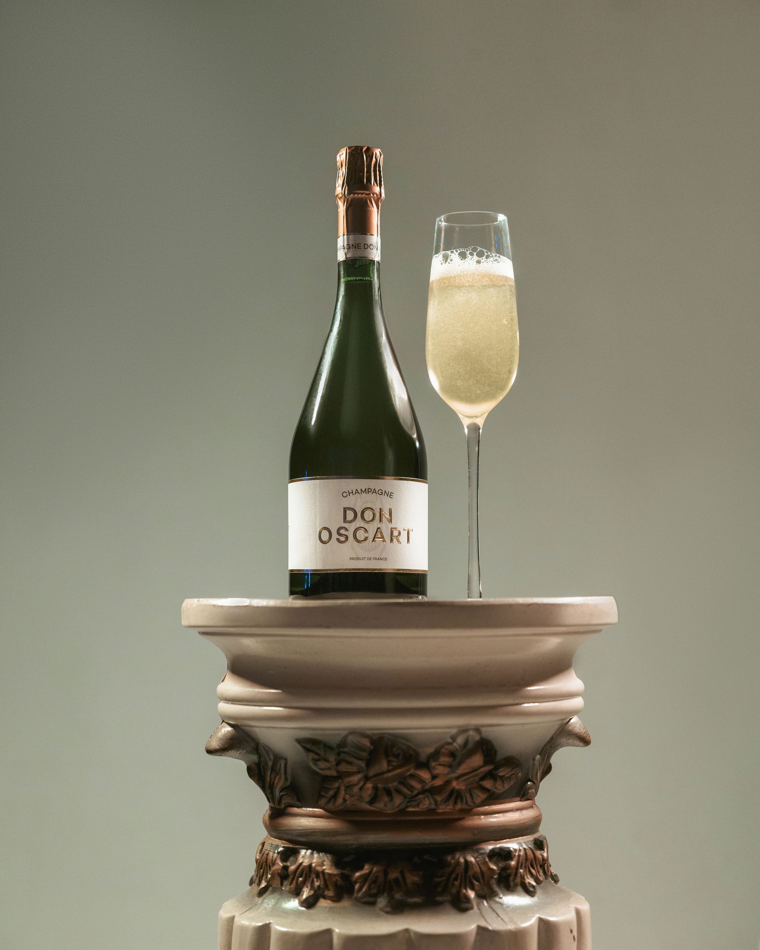 Don Oscar champagne bottle and filled flute displayed on ornate pedestal.